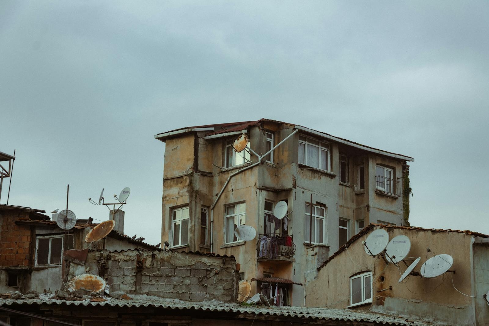 A worn-down residential building with numerous satellite dishes under a cloudy sky.
