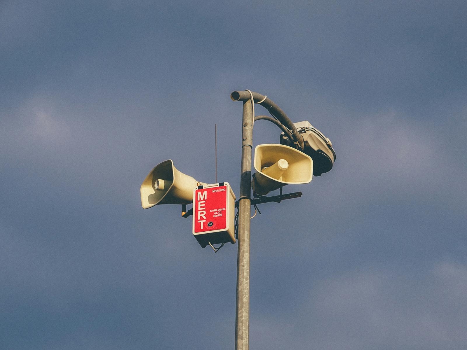 A warning system with loudspeakers on a pole against a cloudy sky, signaling emergency alerts.