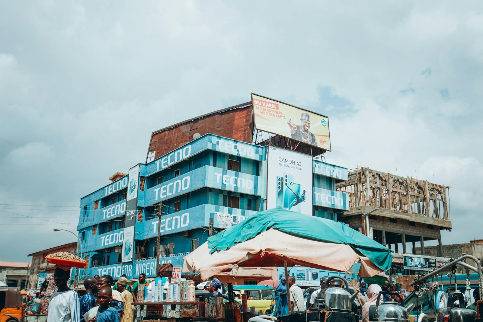 Lively street market scene in Kaduna, Nigeria, featuring vibrant Tecno Mobile advertising and local activity.