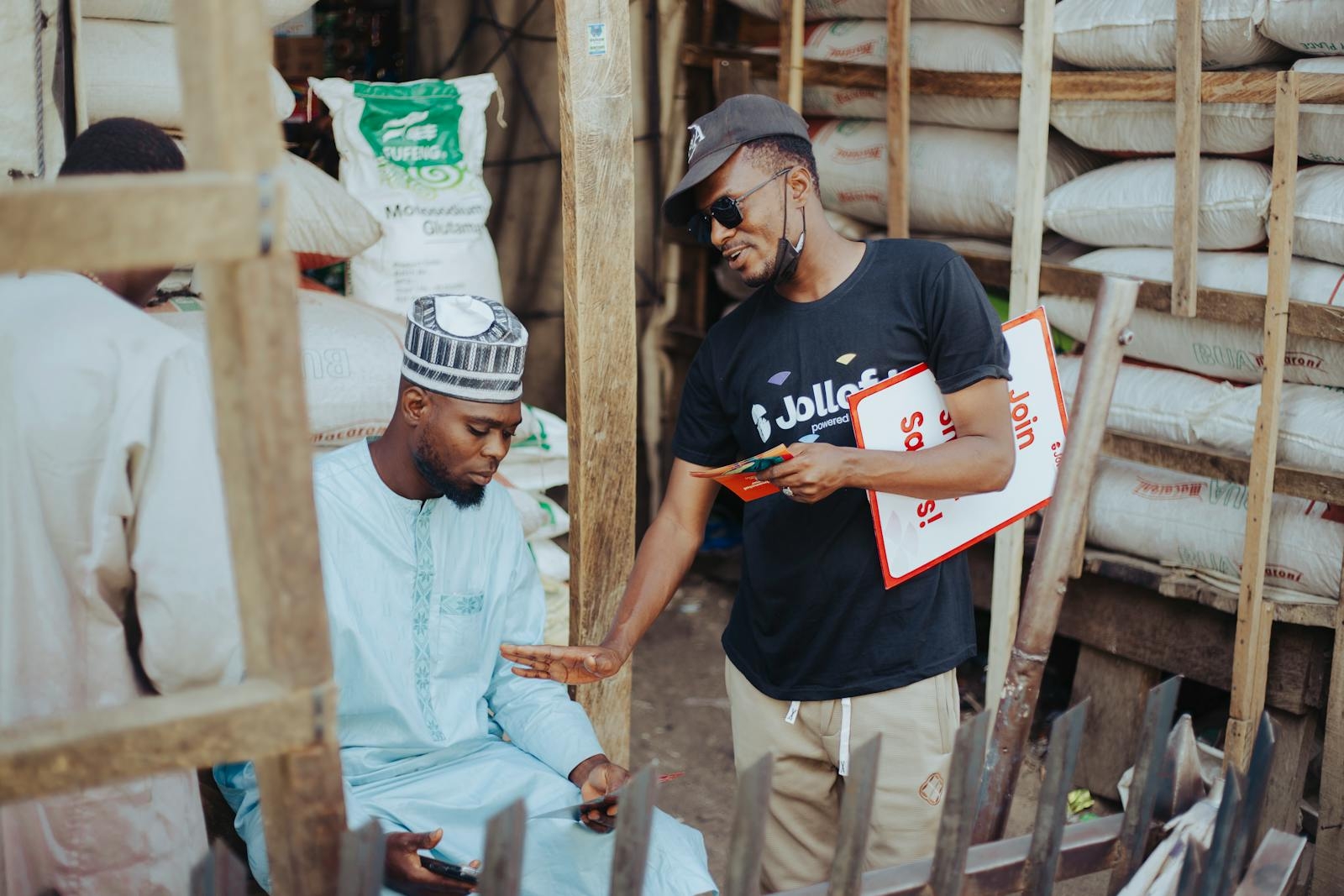 Two men interacting in an outdoor market, surrounded by sacks of goods.