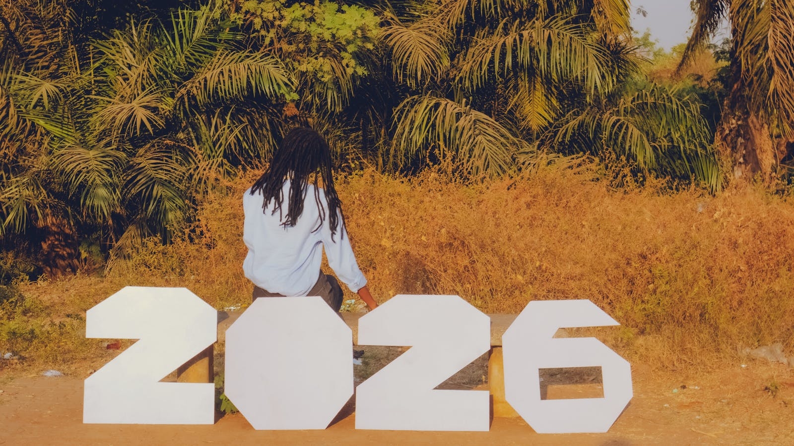 A person sitting outdoors with large numbers spelling 2026, surrounded by tropical foliage.